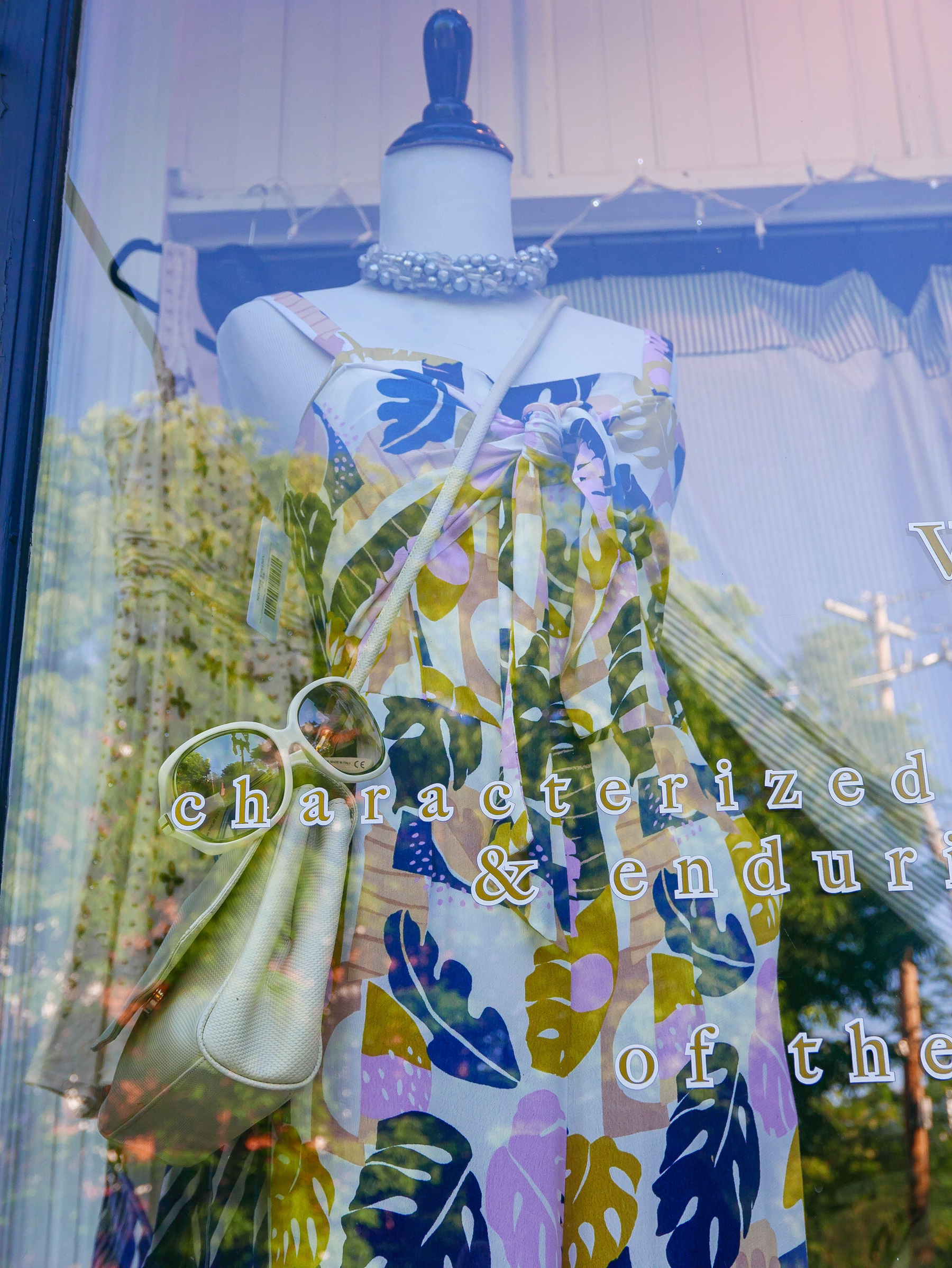 Female mannequin form wearing a tropical leaf print bare shoulder dress gathered at the waist. A purse is slung over the shoulder from right to left. Sunglasses balance precariously on the front edge of the purse.