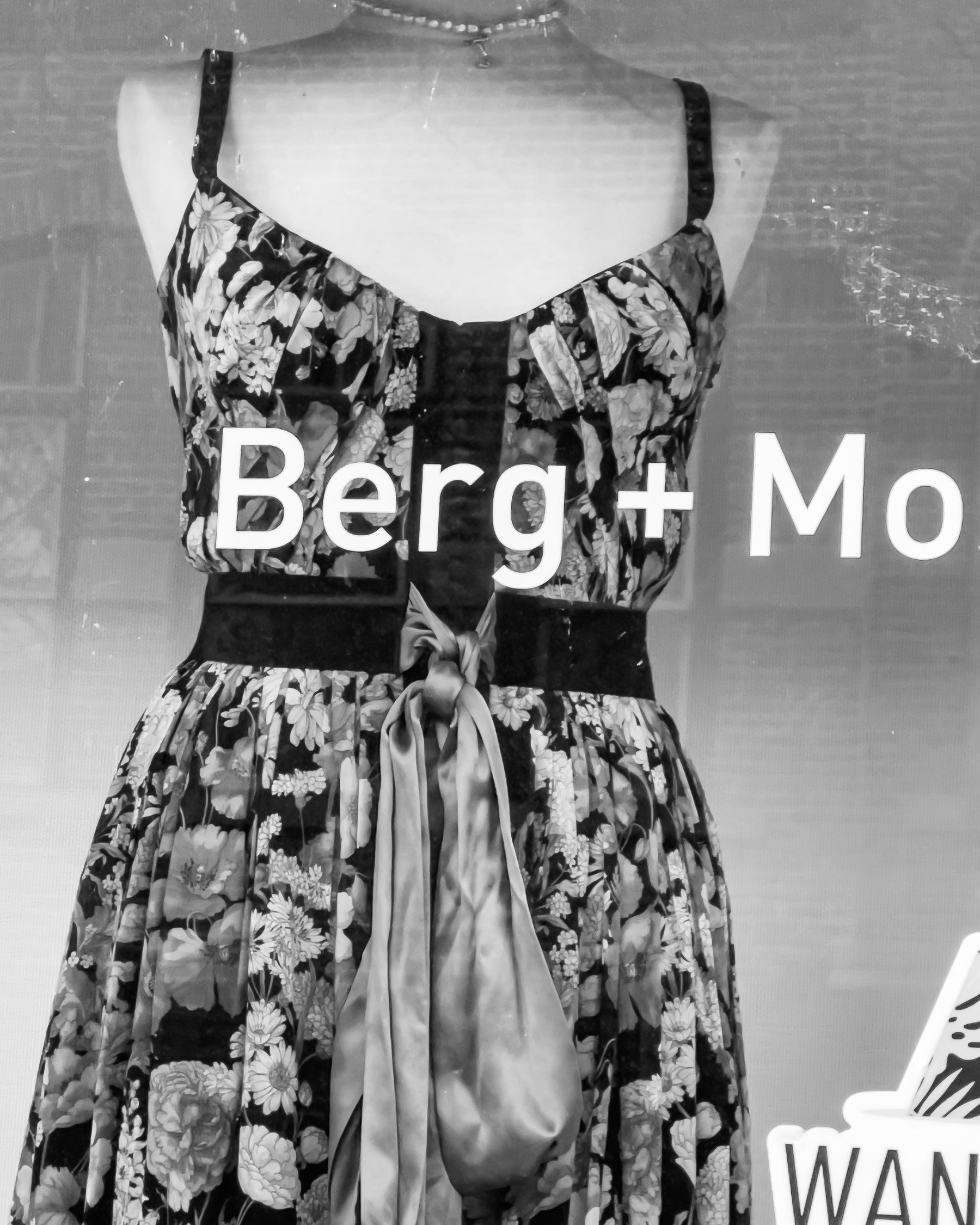 A bare shouldered floral print dress gathered at the waist on a female mannequin form in a shop window. Barg + Mo stenciled on the window just below the breasts of the mannequin form.