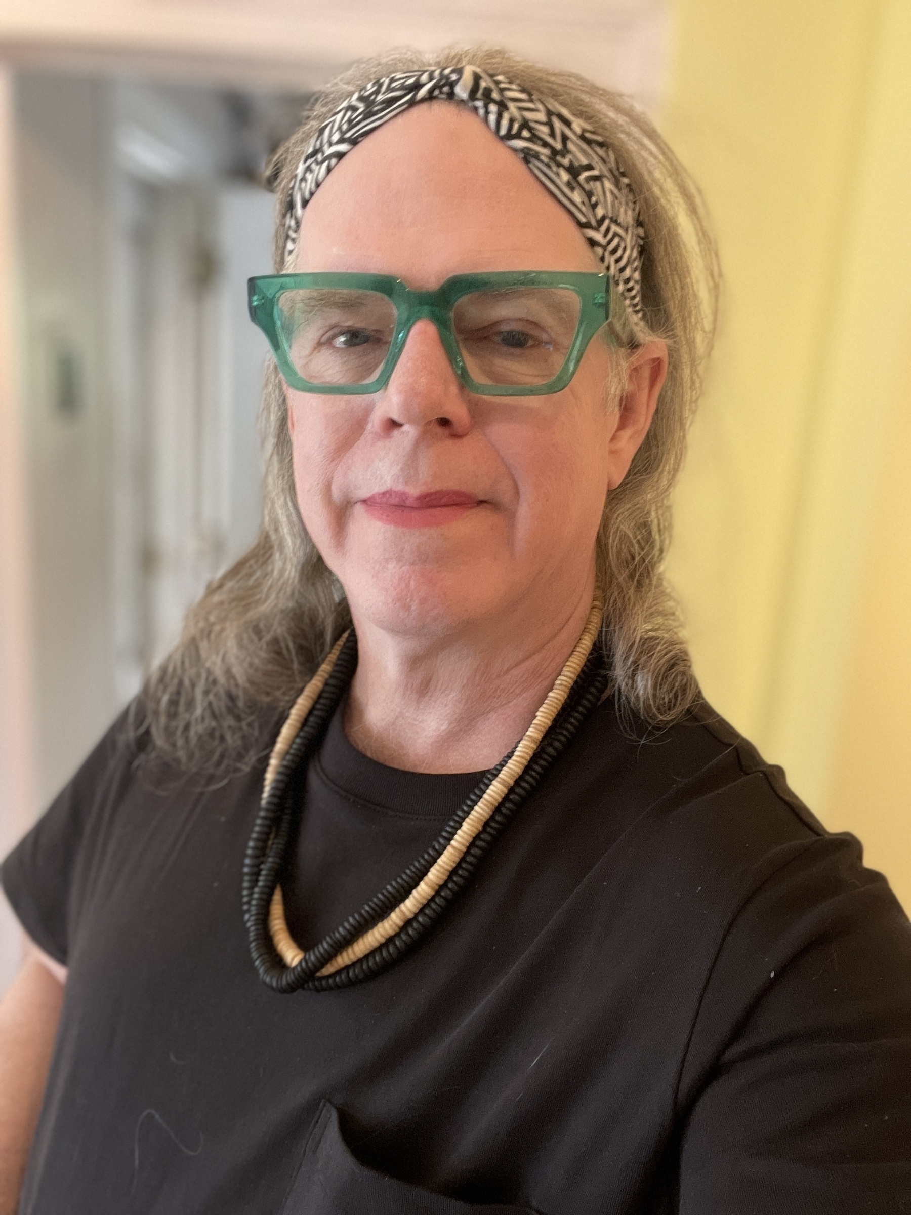 Trans feminine person with wood bead necklace, black cotton top, bold green crystal frame glasses, black and white batik headband, hair cascading in curls to her shoulders and red/pink lipstick. 